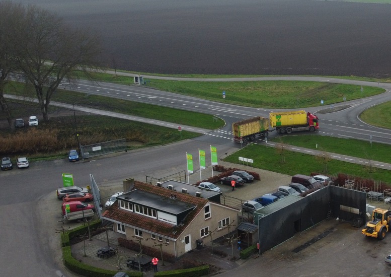 Luchtfoto van de vestiging met kantoorpand, parkeerplaats en een vrachtwagen met gele containers die de weg oprijdt