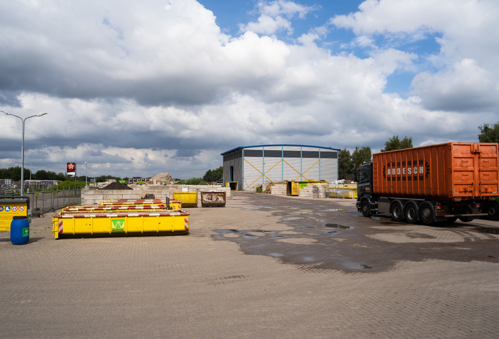Het terrein van Recycling Balkbrug met gele containers, de blauwe loods en een vertrekkende vrachtwagen met oranje container