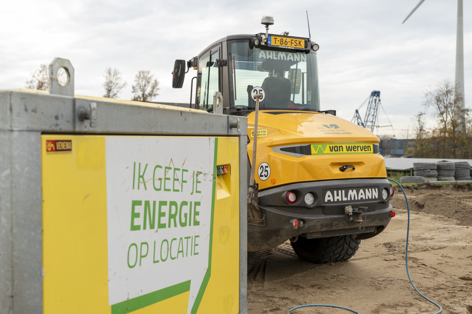 Gele Van Werven-accubox met de tekst 'Ik geef je energie op locatie' laadt via een blauwe kabel een elektrische Ahlmann shovel op, met een windmolen op de achtergrond