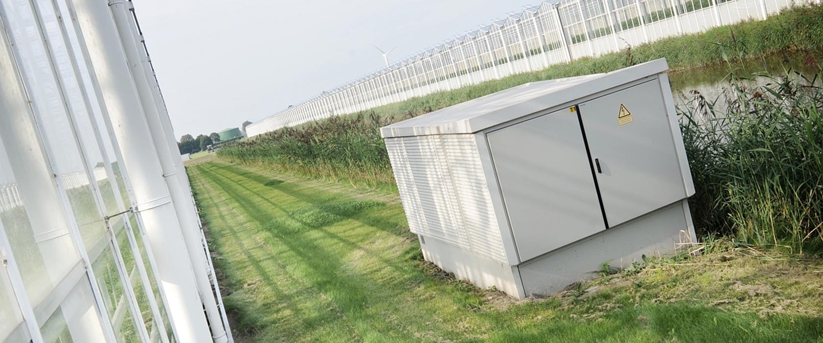Een transformatorhuisje van Alfen in het gras langs een langgerekt kassencomplex