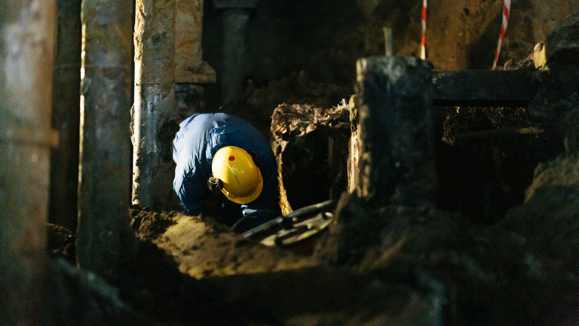 Een medewerker met gele helm graaft gehurkt tussen de oude funderingen, bijgelicht door een bouwlamp