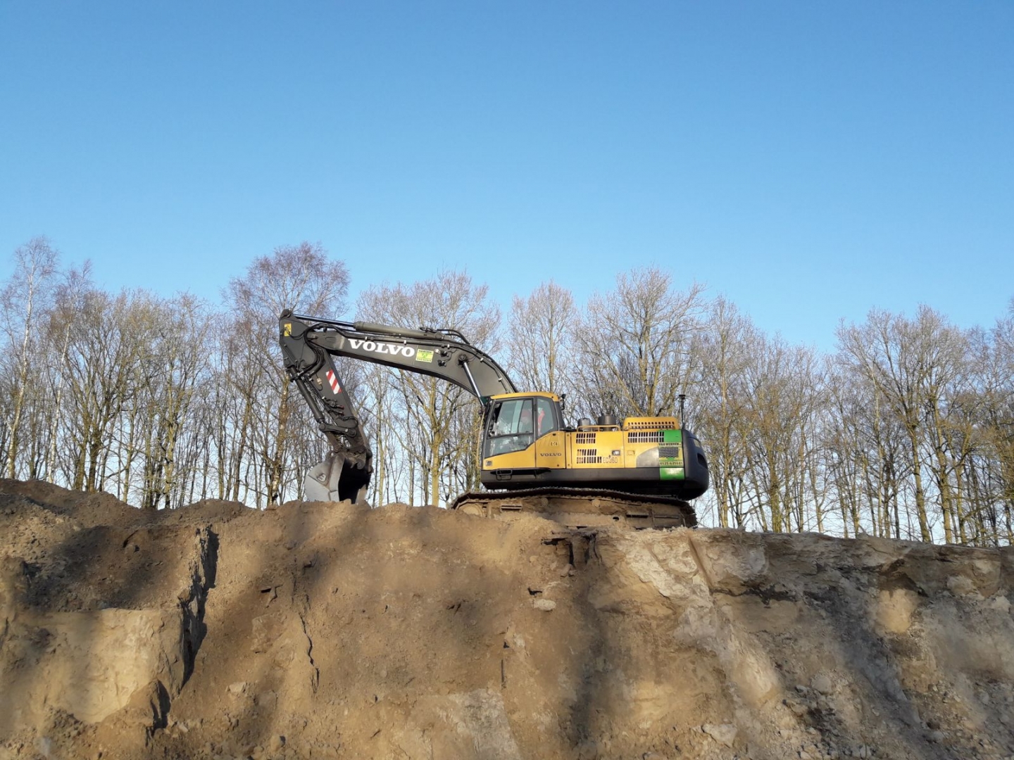 Een Volvo-graafmachine van Van Werven staat op de rand van de uitgraving, tegen een strakblauwe lucht en kale berken