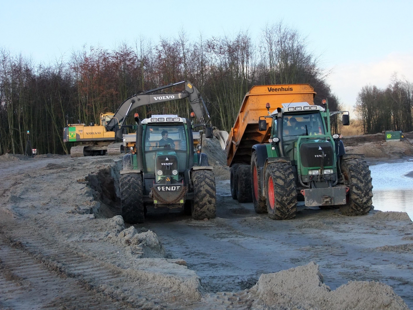 Een Volvo-rupskraan laadt een dumperwagen tussen twee groene Fendt-trekkers op het zandpad