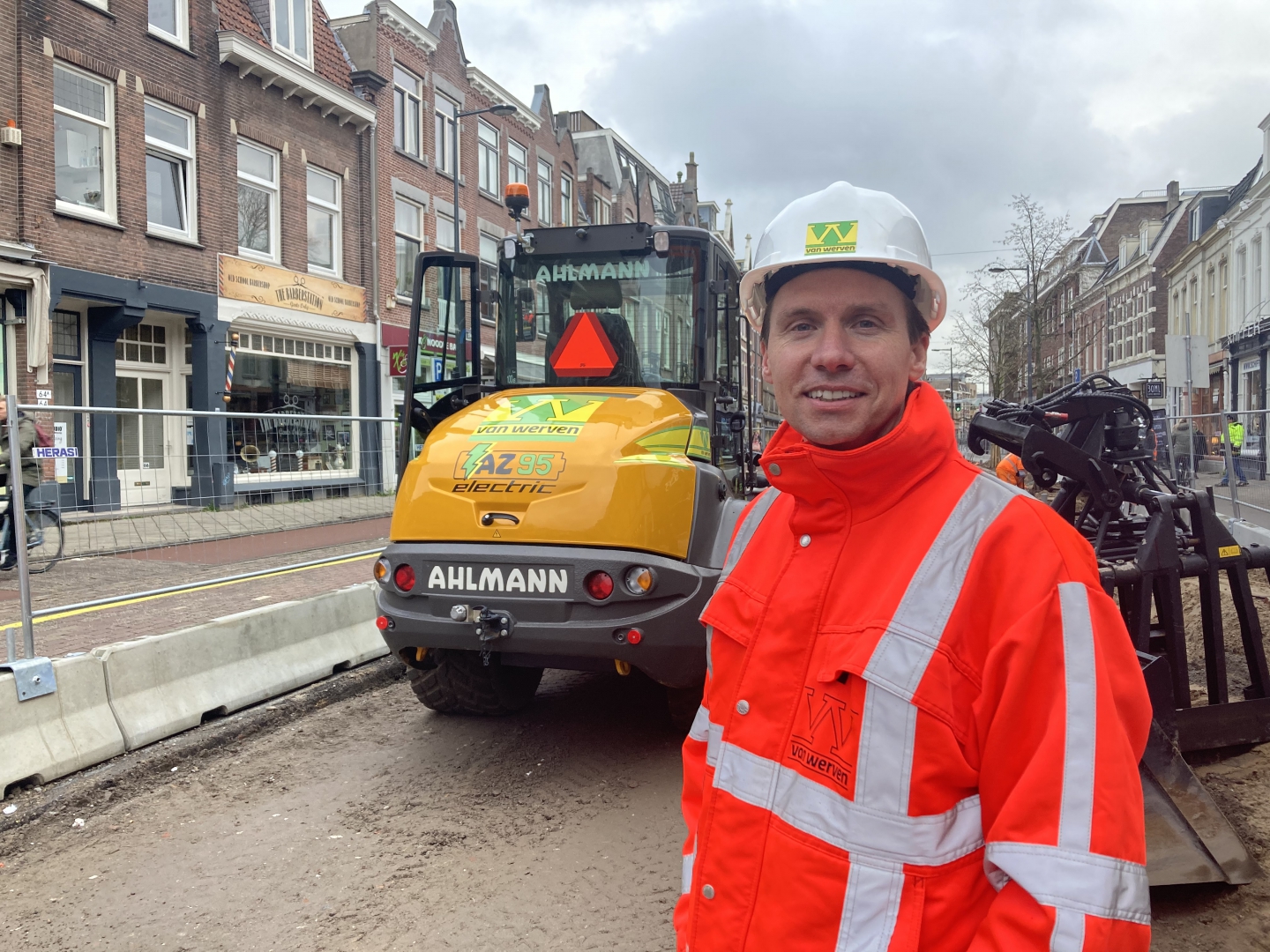 Een medewerker van Van Werven in oranje jas en met witte helm, voor de elektrische Ahlmann-shovel in de winkelstraat