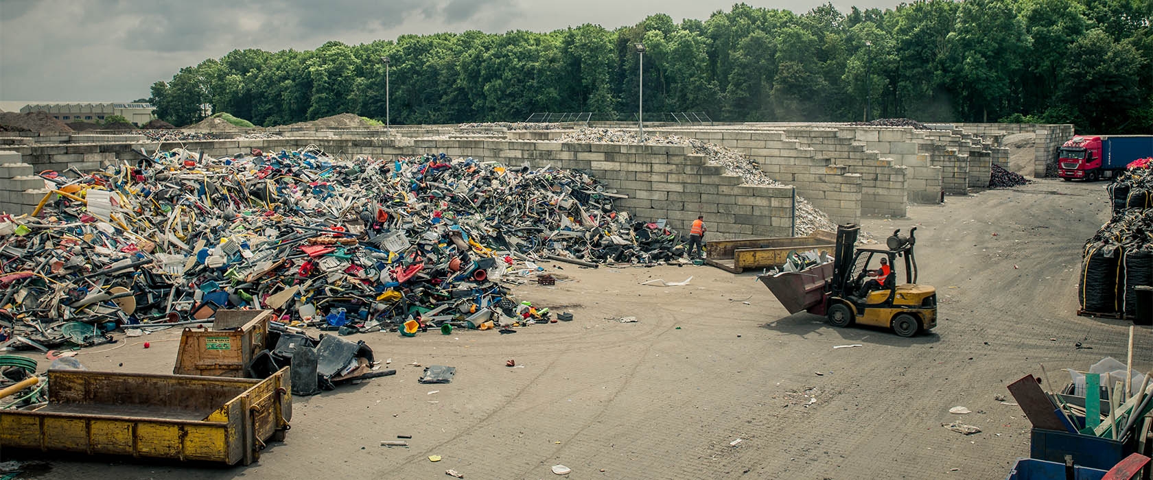 Het buitenterrein van de kunststofrecycling: bergen hard kunststof tussen keerwanden van betonblokken, met een heftruck aan het werk en een vrachtwagen bij de weegbrug