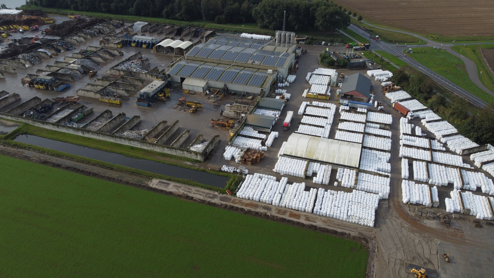 Luchtfoto van de kunststofrecyclinglocatie met honderden witte big bags, zonnepanelen op de daken en betonnen opslagvakken