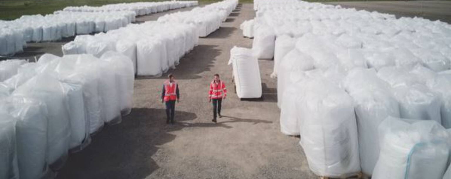 Twee medewerkers in veiligheidshesjes lopen tussen lange rijen witte big bags met plastic