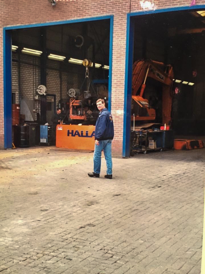 Een jonge medewerker loopt over het erf langs de open werkplaatsdeuren, met een oranje Halla-kraan binnen