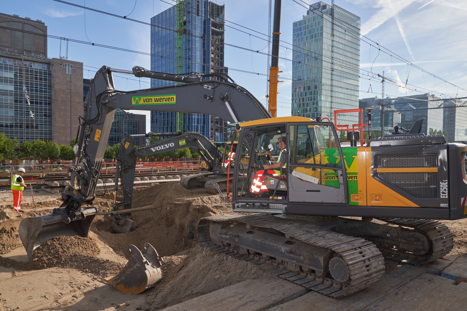 Een Volvo-rupskraan van Van Werven ontgraaft naast de spoorbaan, terwijl een grondwerker met de schop bijsteekt