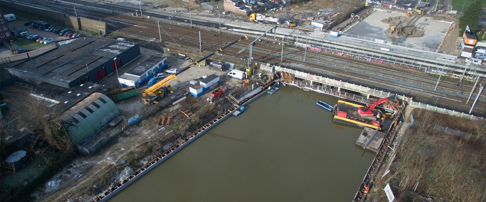 Luchtfoto van een met water gevulde bouwkuip naast het spoor, met materieel langs de rand