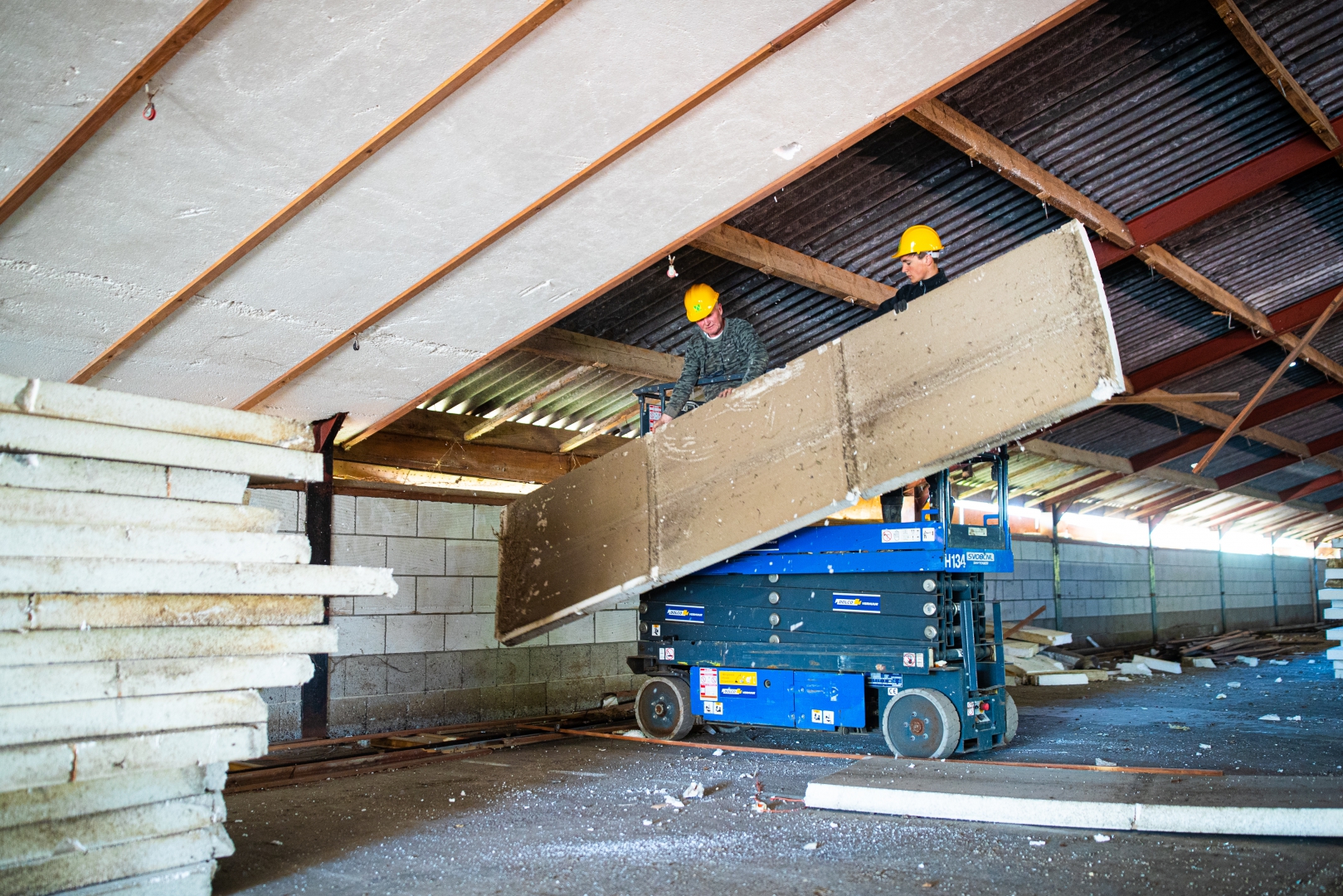 Twee werknemers met helm nemen vanaf een schaarhoogwerker een isolatiedakplaat uit de constructie, met de al verwijderde platen op een stapel links