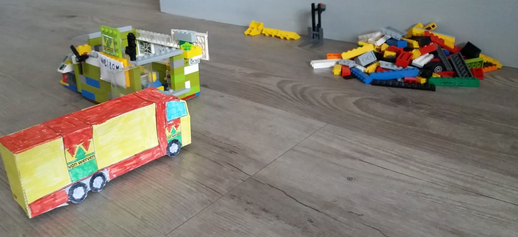 Een zelfgeknutselde Van Werven-vrachtwagen van beschilderd karton op een vloer, naast een bouwwerk en een hoop losse LEGO-blokjes