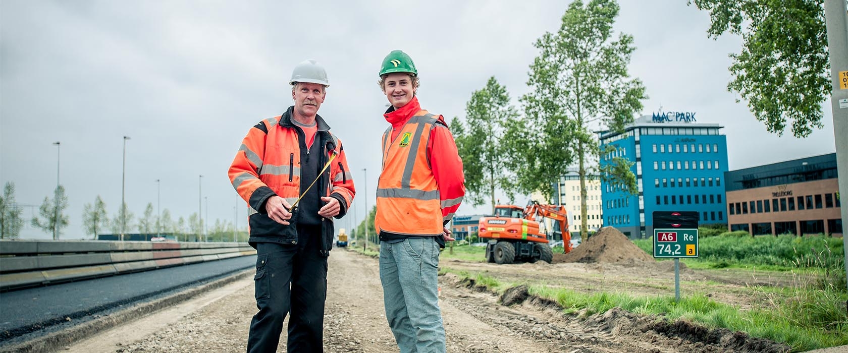 Twee mannen in oranje werkkleding en helm staan op de nieuwe oprit langs de A6, met een hectometerpaal 74,2 en kantoorgebouwen op de achtergrond