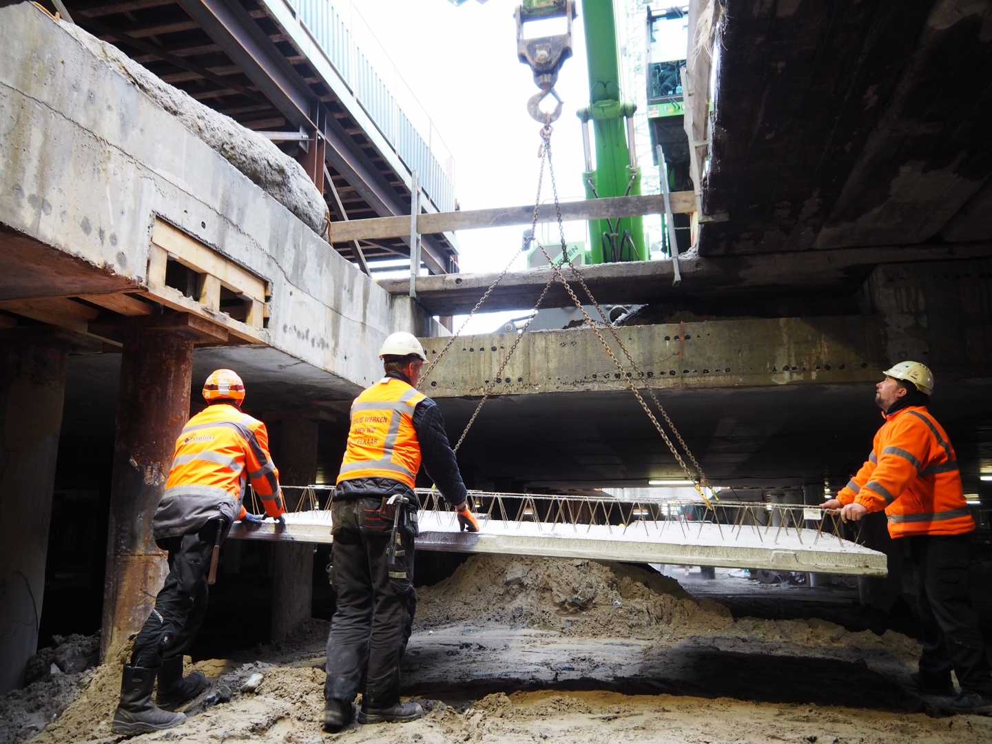 Drie bouwvakkers begeleiden een betonnen breedplaat die aan kettingen onder het bestaande dek wordt gehesen