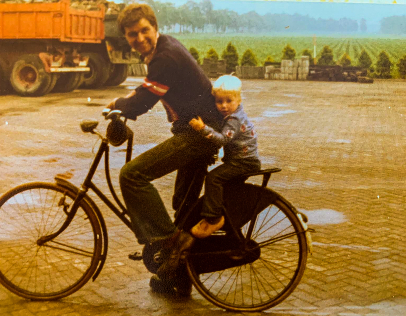 Hendrik op de fiets over het erf, met een blond zoontje achterop en een rode kiepauto op de achtergrond