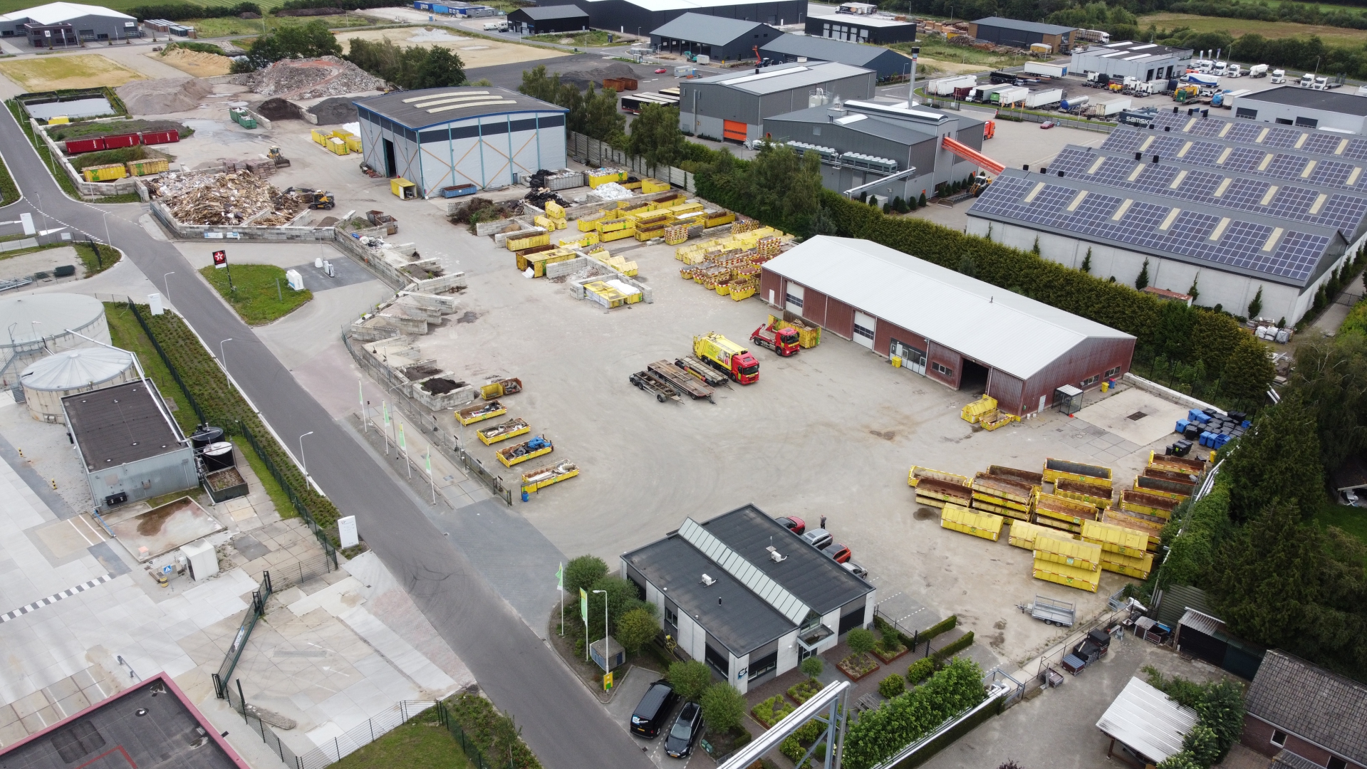 Luchtfoto van de recyclinglocatie met kantoor, rode loods, rijen gele containers en de blauwe hal op de achtergrond