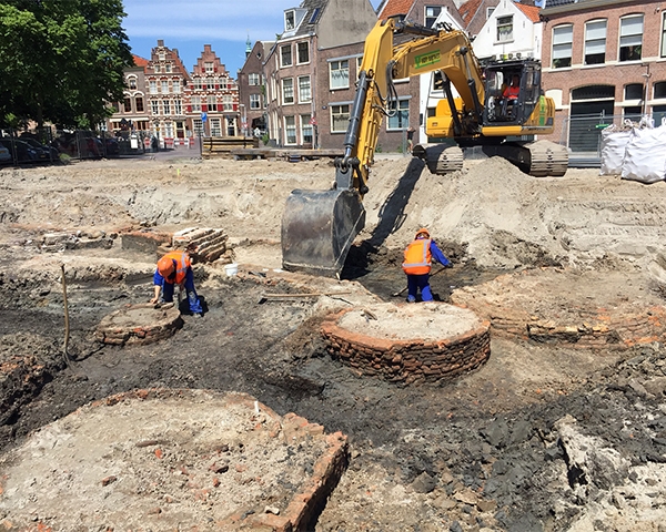 Archeologen in oranje hesjes leggen ronde bakstenen waterputten bloot op de bodem van de ontgraving