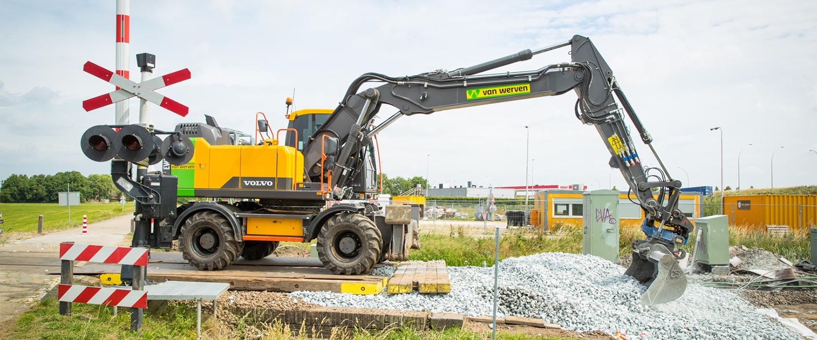 Een Volvo-spoorkraan van Van Werven schept ballast naast een overweg met andreaskruisen