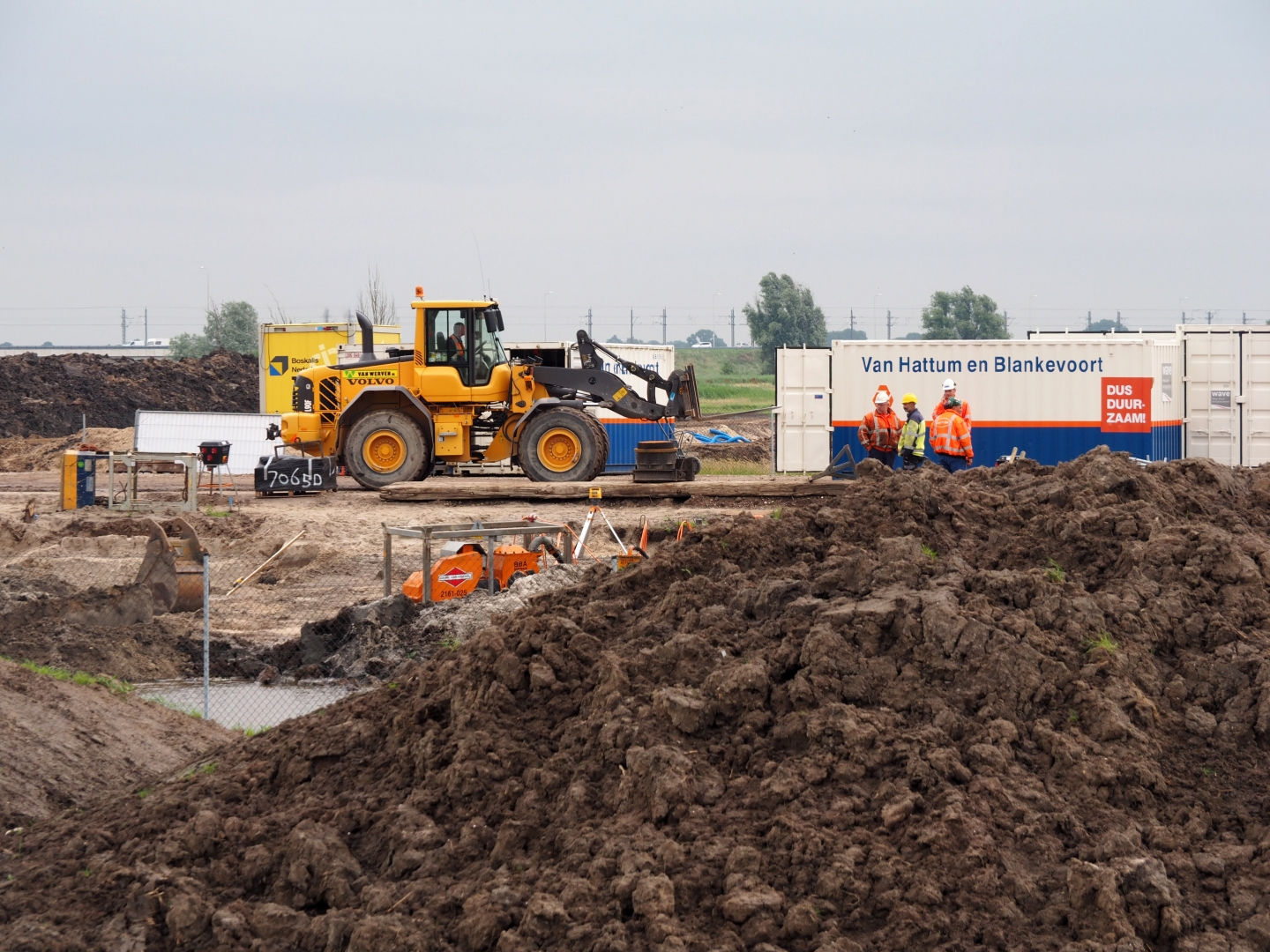 Een gele Volvo-wiellader van Van Werven rijdt over rijplaten langs bouwketen van Van Hattum en Blankevoort, met donkere klei op de voorgrond