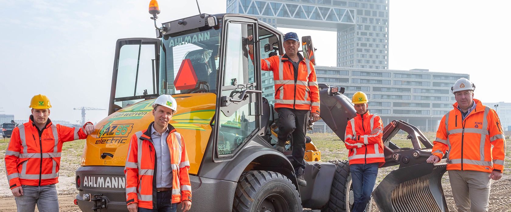 Vijf mannen in oranje werkkleding poseren rond de elektrische Ahlmann AZ 95 shovel op een bouwplaats in Amsterdam