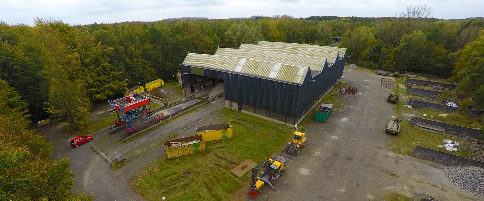 Luchtfoto van de langgerekte zwarte Deltagoot-hal tussen de bomen van het Waterloopbos, met containers en machines op het terrein ervoor
