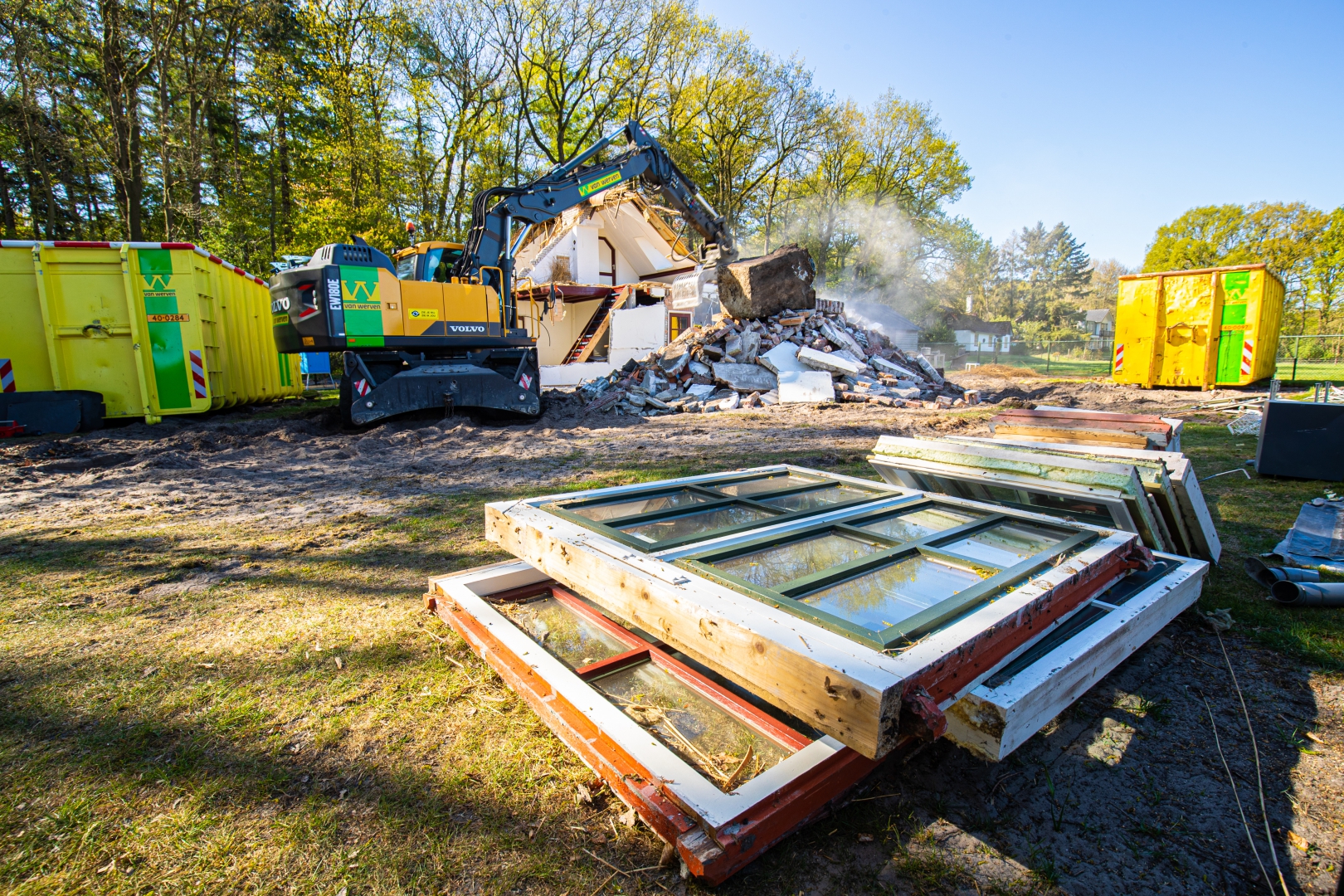 Uitgenomen houten kozijnen met glas liggen op het gras voor hergebruik, terwijl op de achtergrond de sloopkraan het woonhuis afbreekt