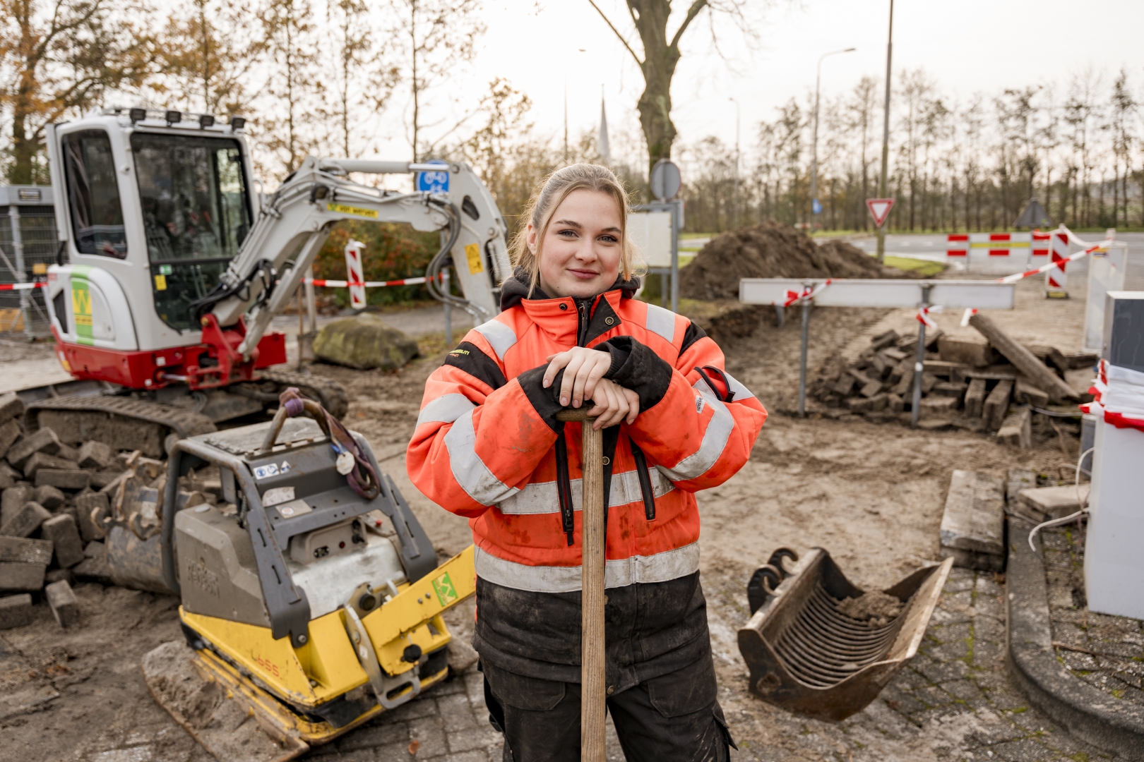 Grondwerker leunt op haar schop bij een bestratingsproject, met minigraver en trilplaat op de achtergrond