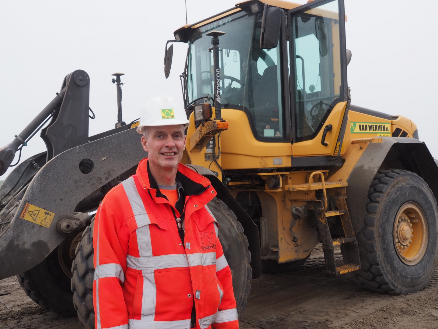 Een medewerker van Van Werven in oranje jas en met witte helm staat glimlachend voor een Volvo-shovel met gps-antennes