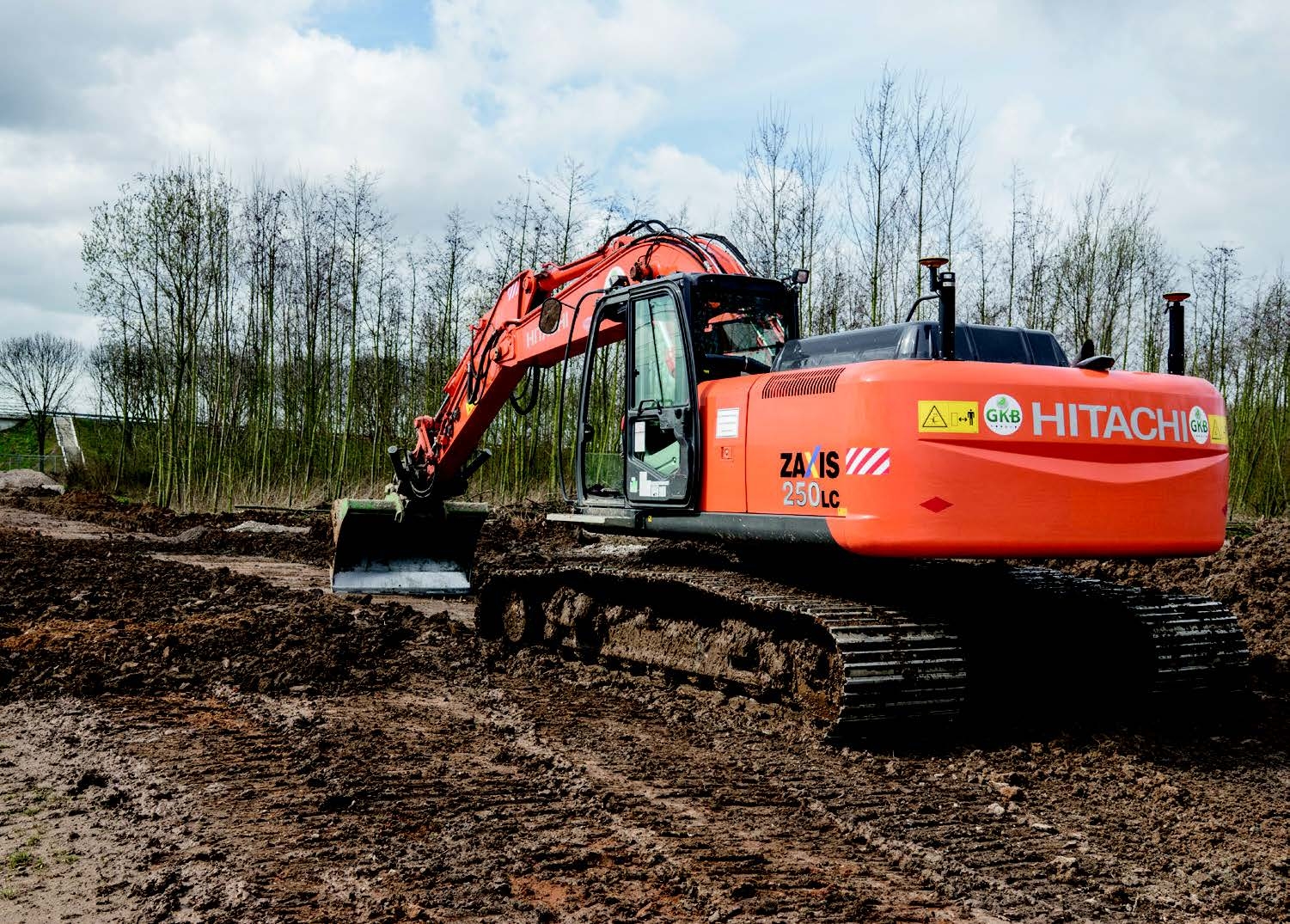 Een oranje Hitachi-rupskraan met twee gps-masten en kilverbak egaliseert een modderig terrein langs jonge aanplant