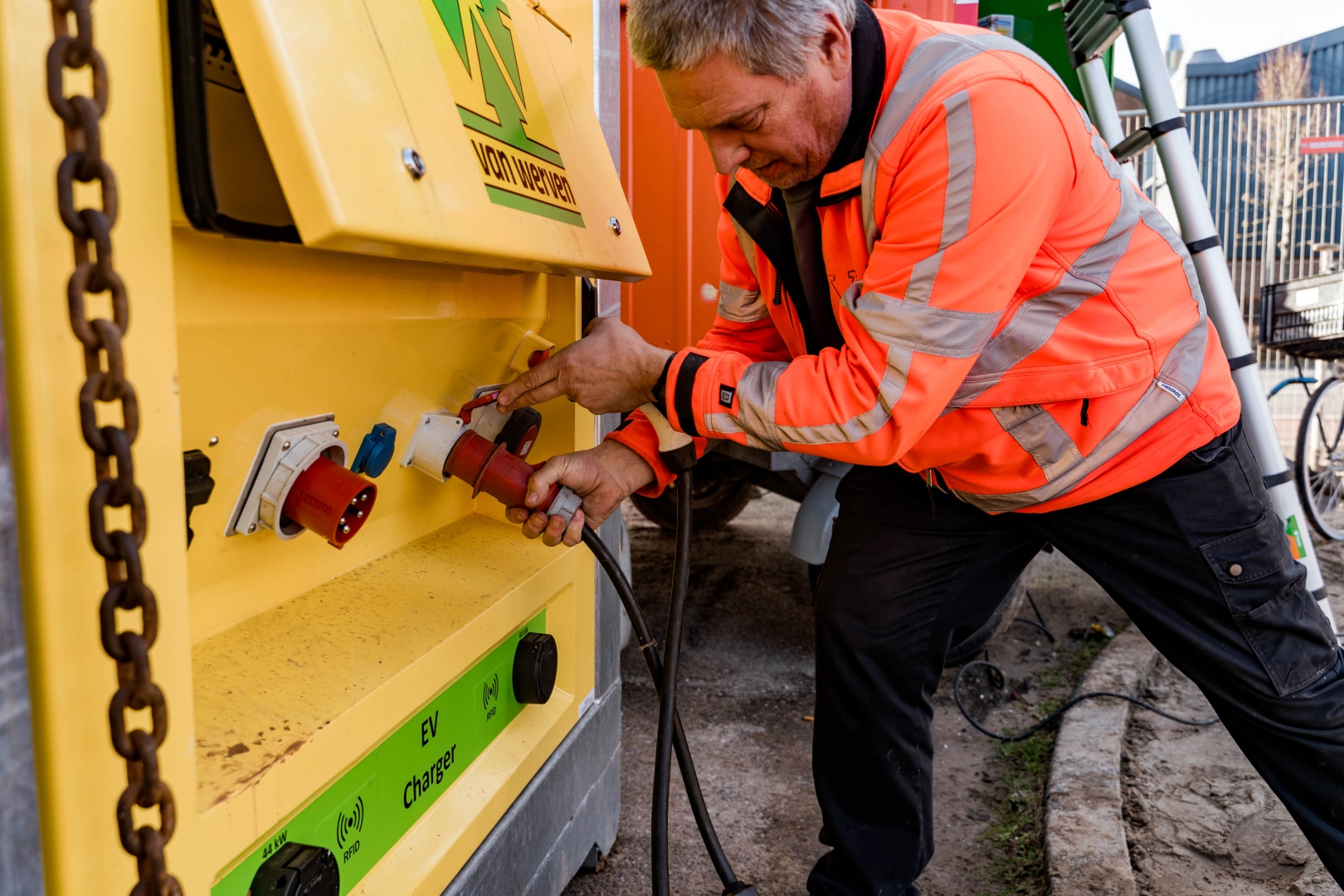 Een medewerker in oranje werkjas steekt een rode krachtstroomstekker in de gele accubox van Van Werven met EV-charger