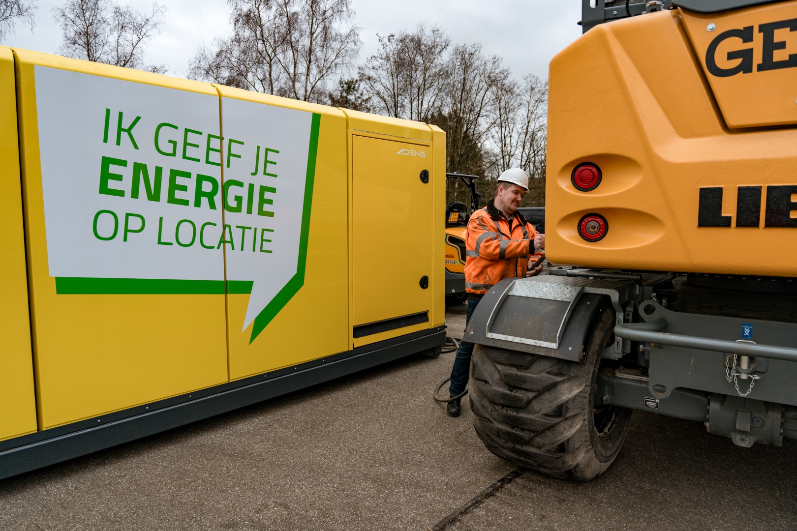 Een medewerker met witte helm sluit een gele Van Werven-accubox met de tekst 'Ik geef je energie op locatie' aan op een Liebherr-kraan