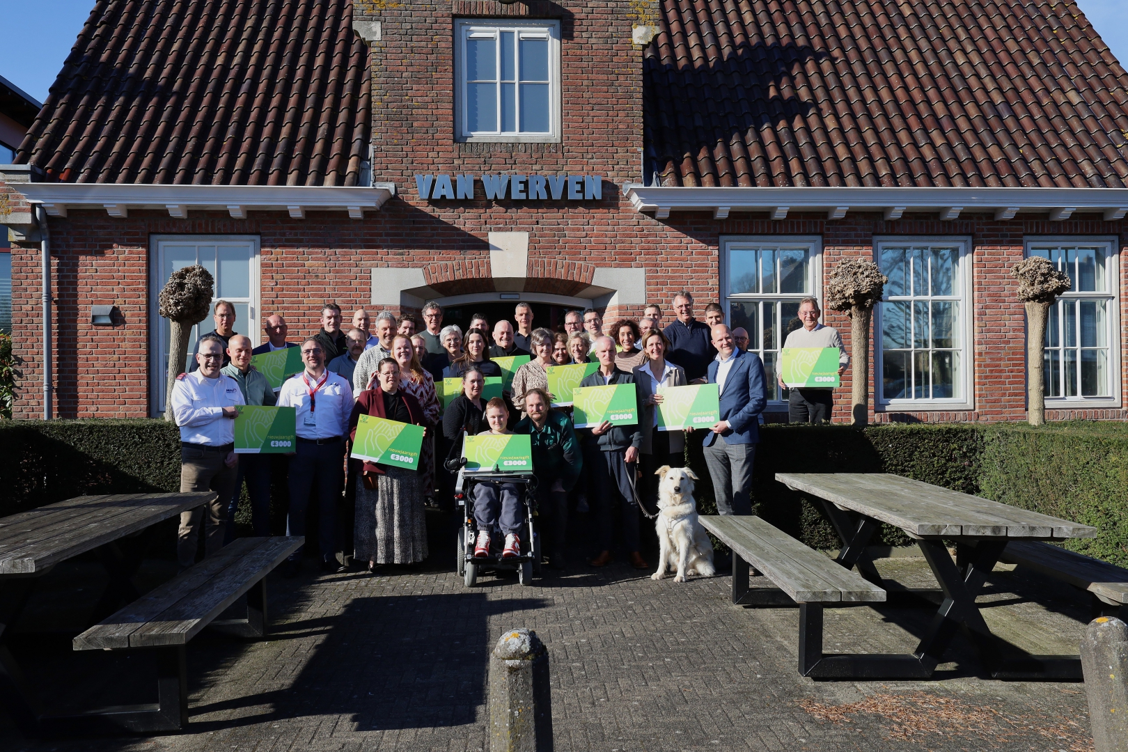 Groepsfoto van vertegenwoordigers van vijftien goede doelen met groene cheques voor het Van Werven-kantoor, met een witte hond vooraan