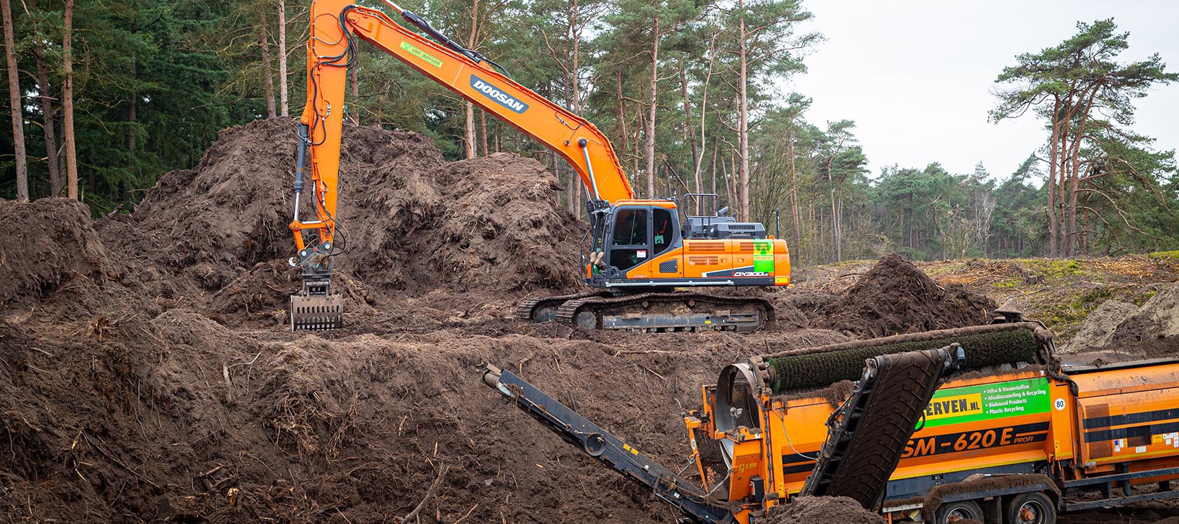 Een Doosan-rupskraan met lange giek en sorteergrijper werkt op de bergen afgegraven bosgrond, voor de trommelzeef