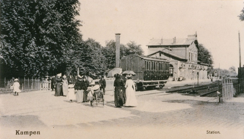 Historische ansichtkaart van station Kampen rond 1900, met een stoomtrein aan het perron en wandelaars op de voorgrond