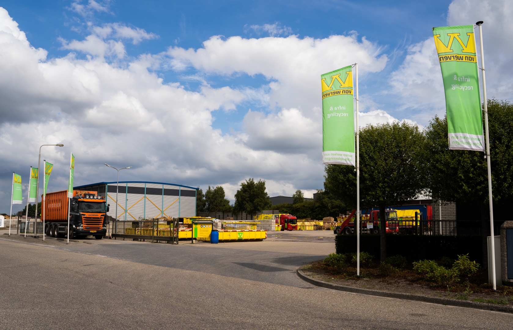 De ingang van Recycling Balkbrug met groene Van Werven-vlaggen, de blauwe loods, gele containers en een vertrekkende vrachtwagen