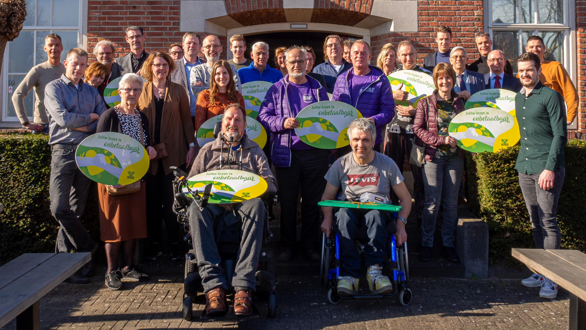 Groepsfoto van vertegenwoordigers van de goede doelen met borden 'Jullie inzet is onbetaalbaar' voor de ingang van het kantoor