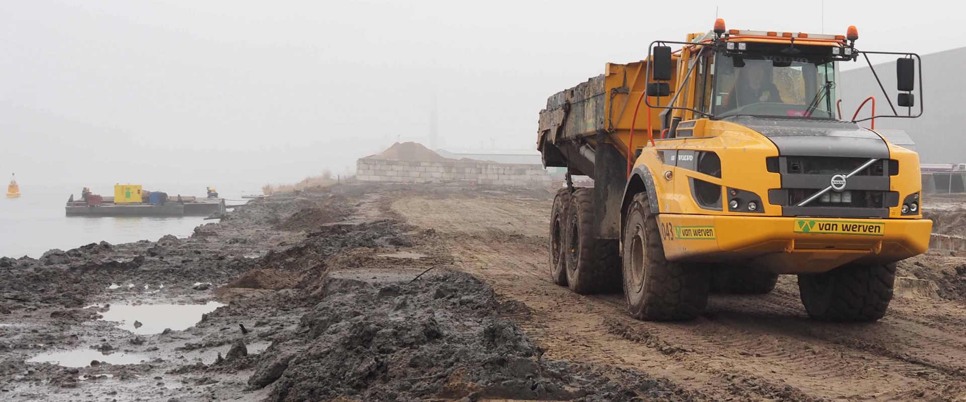 Een Volvo-dumper van Van Werven rijdt over de modderige kade, met het werkponton op het water in de mist