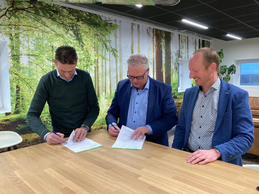 Drie mannen ondertekenen de samenwerkingsovereenkomst aan een houten tafel voor een wandfoto van een bos