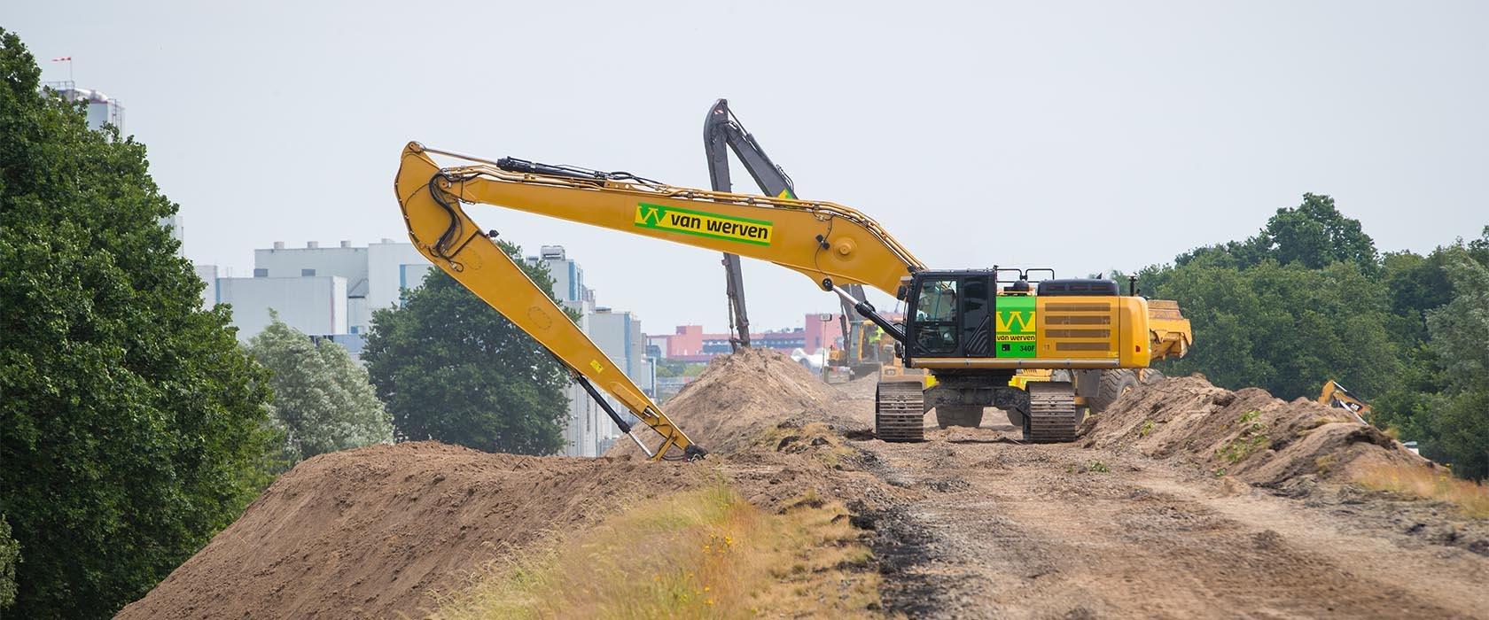 Een rupskraan van Van Werven met lange giek werkt op het zandlichaam langs de spoorbaan, met industrie op de achtergrond