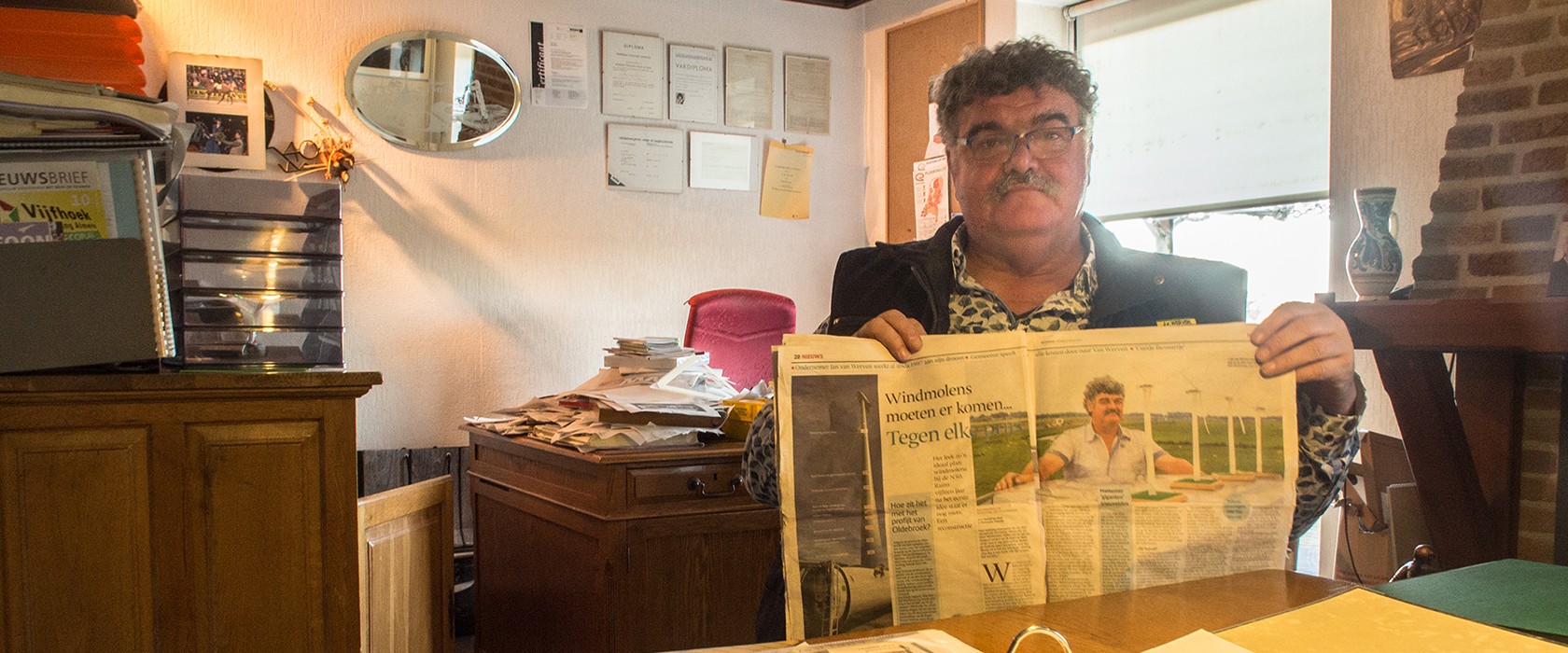 Jan van Werven houdt in zijn kantoor een krantenpagina omhoog met de kop "Windmolens moeten er komen... tegen elke prijs"