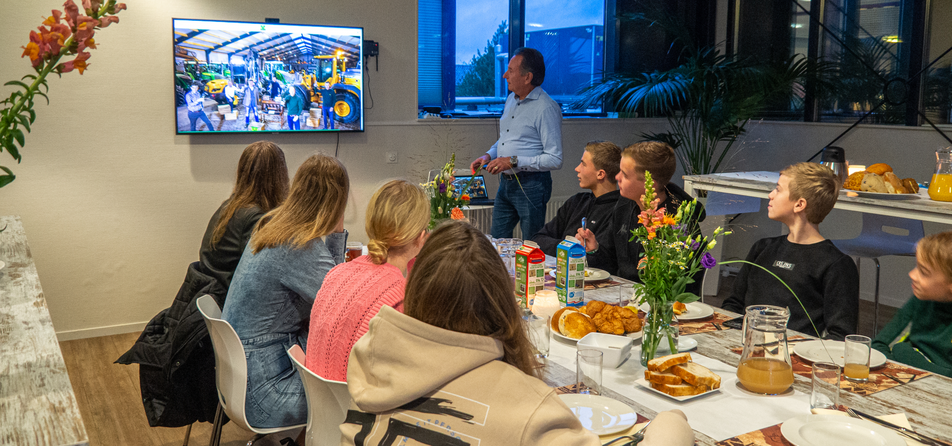 Een man toont een bedrijfsvideo op tv aan een groep jongeren aan een gedekte ontbijttafel