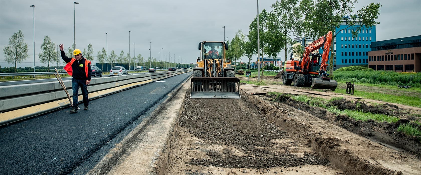 Een wiellader vlakt de puinfundering van de nieuwe rijstrook af terwijl een verkeersregelaar het verkeer op de A6 ernaast stilhoudt