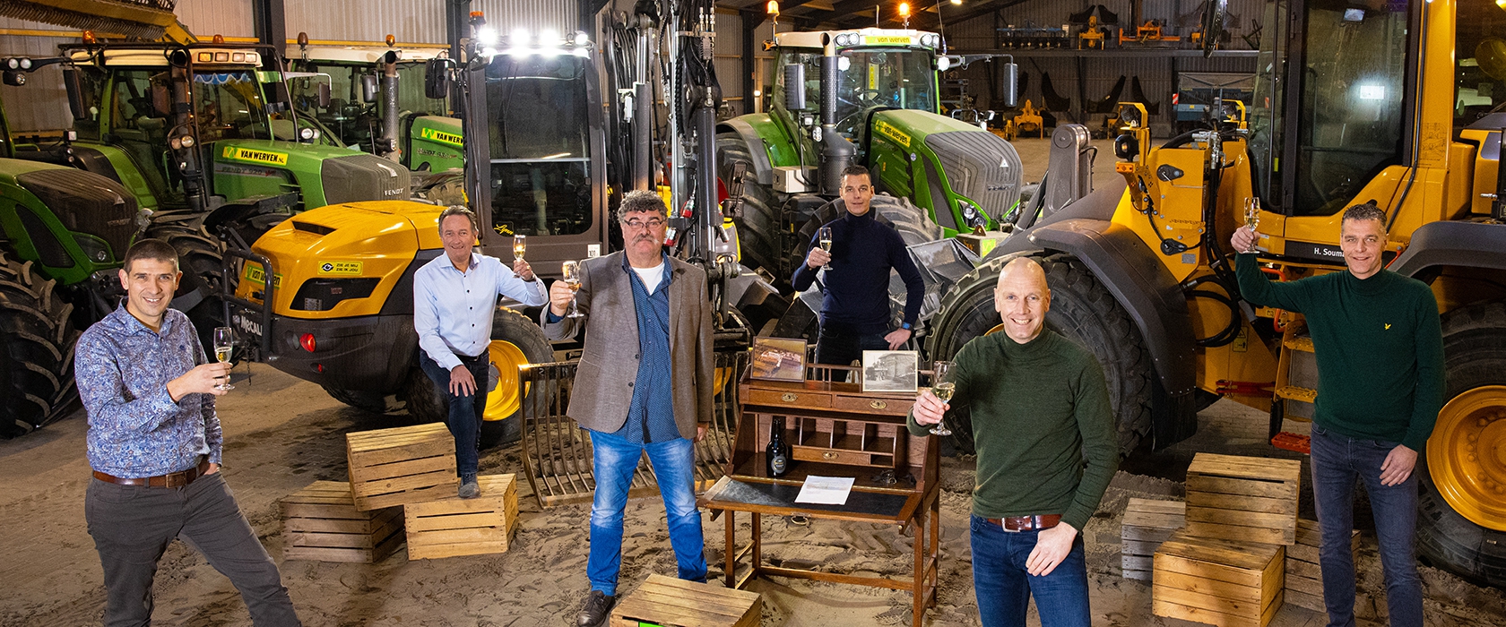 Zeven mannen heffen het glas rond een antiek bureau in de loods, tussen tractoren en ander materieel