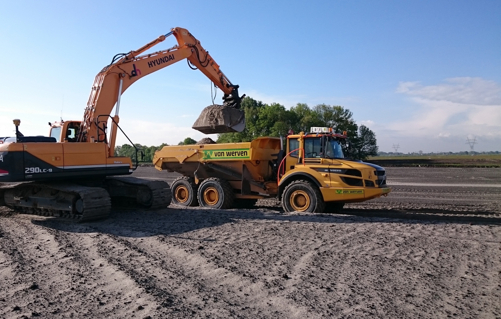 Een Hyundai-graafmachine laadt grond in een gele Van Werven-dumper op het vlakke werkterrein