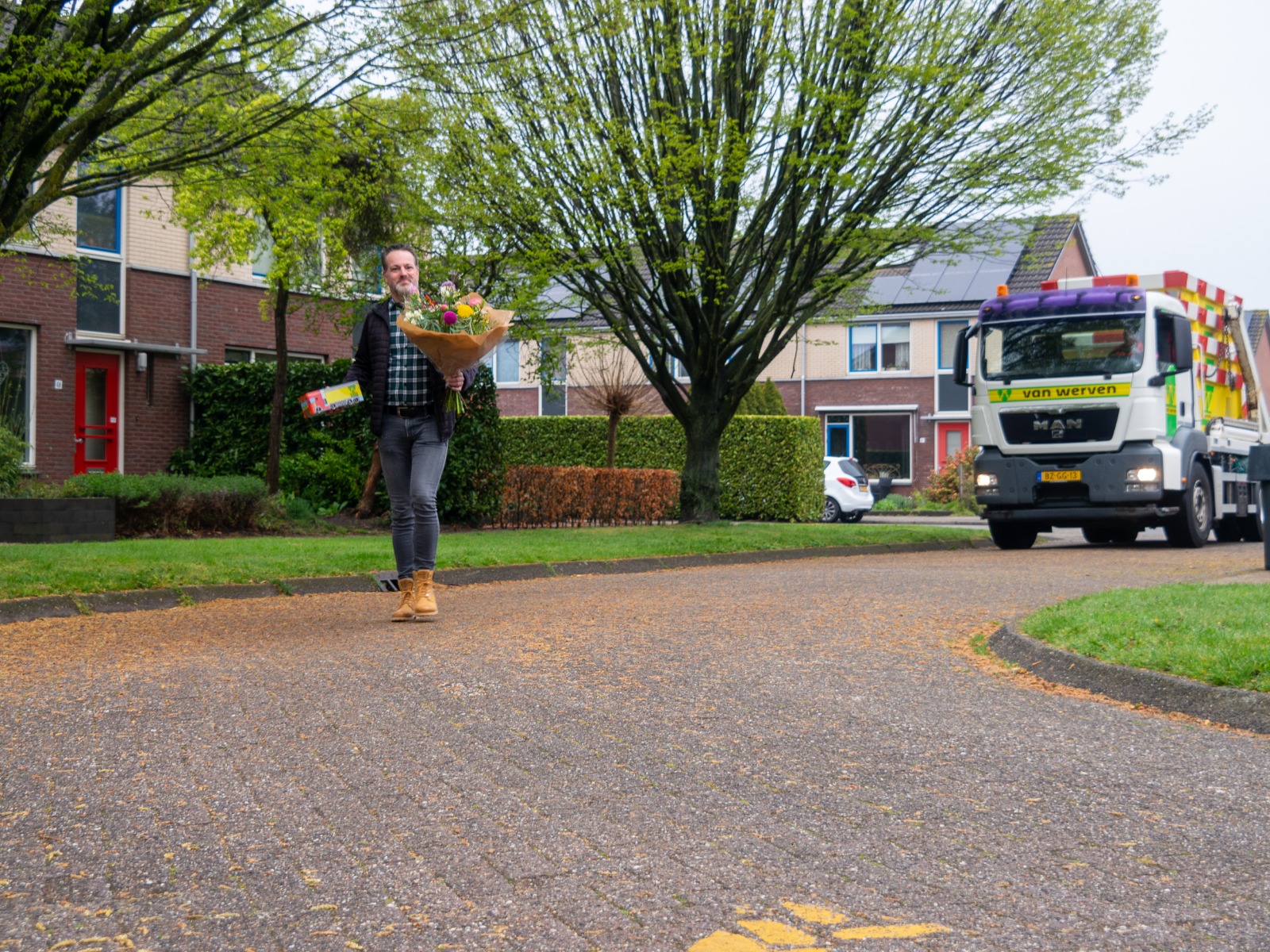 Een man brengt een bloemboeket en een cadeau door de woonwijk; op de achtergrond staat de portaalarmwagen van Van Werven