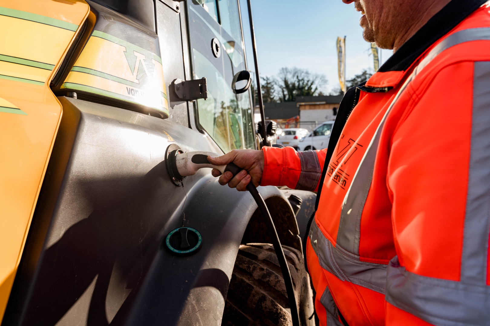 Een medewerker in oranje werkjas steekt de laadstekker in de elektrische shovel van Van Werven