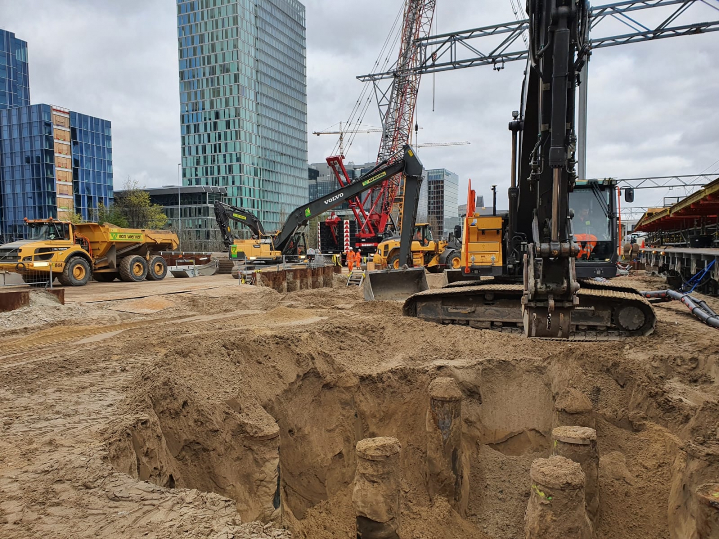 De ontgraven bouwput met de koppen van oude houten funderingspalen, tussen een dumper en een rupskraan, met de torens van de Zuidas erachter