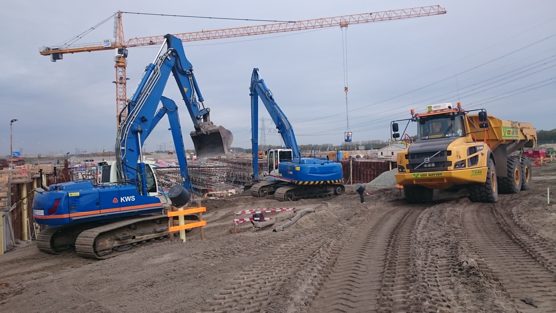 Twee blauwe rupskranen van KWS en een gele Van Werven-dumper bij een diepe bouwkuip, onder de arm van een torenkraan