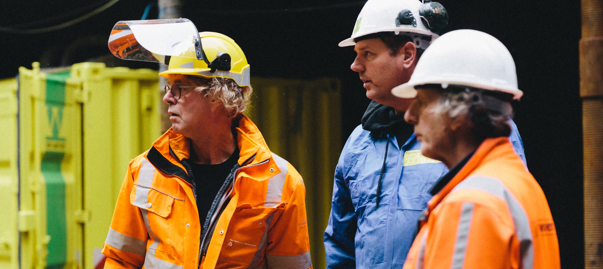Drie mannen met helm, van wie één met gelaatsscherm, in overleg voor de geel-groene containers van Van Werven