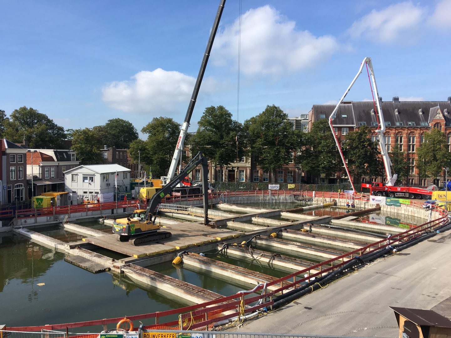 De volgelopen bouwkuip als bassin: een rupskraan met lange giek werkt vanaf het dek tussen de betonnen balken, naast de betonpomp